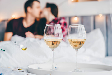 Wall Mural - Glasses of white wine in the foreground and out of focus happy young man and woman kiss on bed in light hotel room closeup.