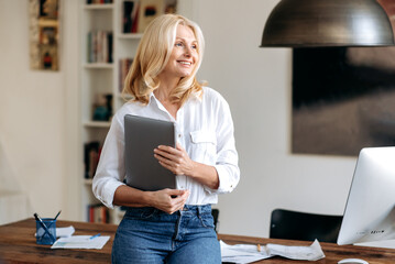 Portrait of a charming elegant caucasian blonde woman, standing in an office near her workplace, dressed in stylish clothes, with a laptop in her hand, looking to the side and smiling friendly