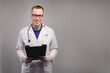 © yanik88 - Portrait of an attractive young Caucasian doctor in glasses and a white coat stands with a tablet in his hands smiling and looking at the camera on a gray background. Studio portrait copy space