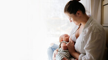 Cute Caucasian Mom And Newborn Baby, Mother Breastfeeding Baby By The Bed In A Bright White Bedroom. Top View, Toning And Lifestyle In A Real Interior