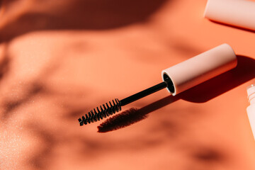 pink clean makeup brush for mascara lies next to an open tube, closed tubes of cosmetics, lip gloss, liquid lipstick, pink eyeliner on a peach background with shadows. Copy space