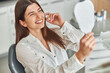 © Ivan - Happy young woman smiling checking out her perfect healthy teeth in the mirror, sitting in a dental chair at the dentist office