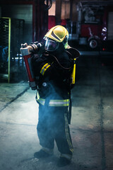 Wall Mural - Portrait of a female firefighter wearing a helmet and all safety equipment a while holding a tomahawk and wearing an oxygen mask indoors surrounded by smoke