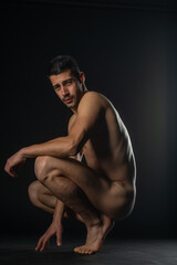 Portrait of a naked male model posing squatted on black studio background