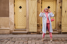 Happy Smiling Fashionable Woman Wearing Trendy Sport Chic Style Outfit Posing In Street Of European City. Blue Trench Coat, Sunglasses, Pink Hoodie, Trousers, Sneakers, Mini Baguette Bag. Copy Space