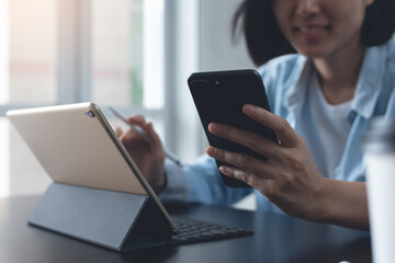 Asian business woman video calling via mobile phone, working on laptop computer on table at home office