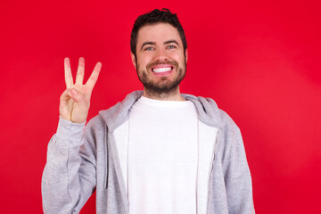 Sticker - young handsome caucasian man in sports clothes against red wall showing and pointing up with fingers number three while smiling confident and happy.