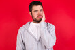 © Jihan - Sad lonely young handsome caucasian man in sports clothes against red wall touches cheek with hand bites lower lip and gazes with displeasure. Bad emotions