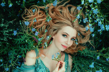 Art Photo Fairy Tale Beauty. Portrait Fantasy Woman Lies On Blooming Meadow In Blue Vintage Dress. Summer Nature Background Spring Green Grass Flowers. Girl Princess Blond Long Hair. Look At Camera