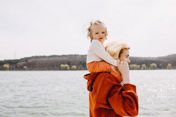 Wall Mural - Mother holding little daughter on shoulders, spending family time together at a lake. Piggyback riding.