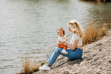 Wall Mural - Young mother playing with her daughter outdoors, sitting by the lake.