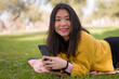 © TheVisualsYouNeed - lifestyle portrait of young happy and beautiful Asian Chinese woman in yellow sweater enjoying with mobile phone cheerful at city green park next to tree in Spring