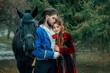 Medieval couple in love man and woman hugging in winter forest. Vintage clothing red long dress. Blue costume tailcoat caftan. Prince and princess together. Black horse. Art image redhead hairstyle.