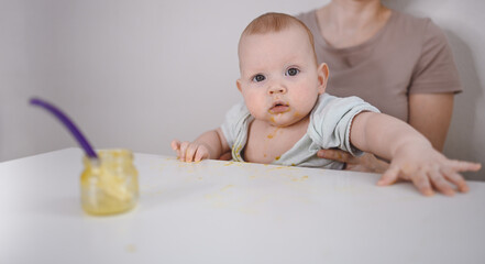 Little newborn funny baby boy learning to eat vegetable or fruit puree from glass jar with spoon. Young mother helping little son eat first food