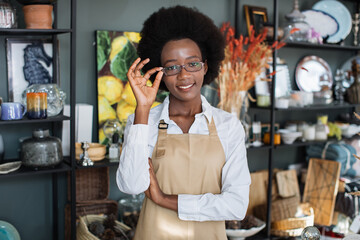 Wall Mural - Waist up portrait of pretty afro american woman in uniform and eyeglasses smiling and looking at camera. Modern shop with various decor on shelves.