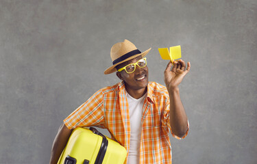 Wall Mural - Studio portrait of happy man ready for flight. Black guy in glasses, orange shirt and summer hat holding yellow travel suitcase and paper airplane smiling at camera isolated on gray background