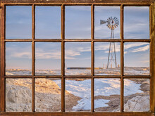 Windmill With A Pump And Cattle Water Tank In A Prairie, Winter Or Early Spring Scenery As Seen From A Vintage Sash Window