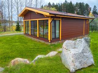 Wall Mural - Country house with a green lawn. Wooden house with panoramic windows. Cottage with panoramic windows. Landscape of the site is decorated with boulders. One-storey cottage by the river.