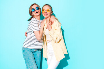 Wall Mural - Two young beautiful blond smiling hipster female in trendy summer clothes. Sexy carefree women posing near blue wall in studio. Trendy and positive models having fun in sunglasses