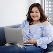 © Bangkok Click Studio - Asian young happy beautiful plus size sexy fat chubby smart female model sit relaxing on sofa smiling look at camera using laptop computer work from home in quarantine time from covid pandemic moment