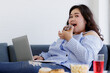 © Bangkok Click Studio - Asian young happy beautiful fat female shopper sit relaxing on fabric sofa look at camera using laptop computer searching products shopping online while eating delicious chocolate donut in hand