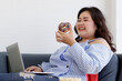 © Bangkok Click Studio - Asian young happy beautiful fat female shopper sit relaxing on fabric sofa look at camera using laptop computer searching products shopping online while eating delicious chocolate donut in hand