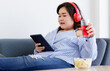 © Bangkok Click Studio - Asian young happy pretty friendly fat plus size woman sit smiling relaxing on gray fabric couch listening to music from big red headphones use playlist from black tablet on hand in living room
