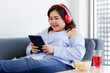 © Bangkok Click Studio - Asian young happy pretty friendly fat plus size woman sit smiling relaxing on gray fabric couch listening to music from big red headphones use playlist from black tablet on hand in living room