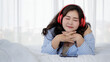 © Bangkok Click Studio - Asian young happy calm fat overweight woman lay down close eyes smiling peacefully on white sheet blanket listening to music from big red earphones use playlist from black tablet at home in bedroom