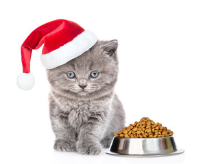 Poster - Cute kitten wearing red santa hat sits with bowl of dry food for pets. isolated on white background