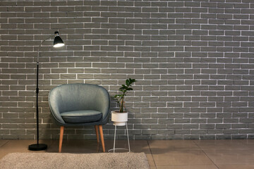 Comfortable armchair with glowing lamp and houseplant near brick wall in room