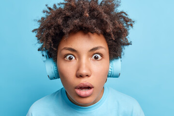 Wall Mural - Close up shot of dark skinned girl with Afro hair stares bugged eyes has startled expression being shocked by something listens music via wireless headphones isolated over blue background. Omg concept