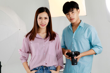 Portrait of two handsome man and beautiful woman wearing casual shirts, holding camera, posing and standing in indoor photo studio with blur background of lighting equipments.