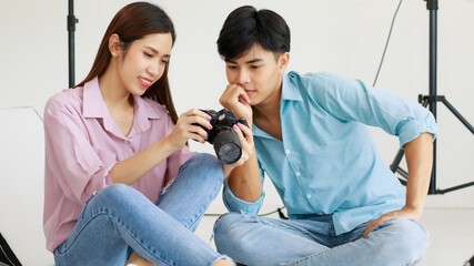 Two handsome man and beautiful woman wearing casual shirts smiling and looking or checking pictures in camera while sitting in indoor photo studio with blur background of lighting equipments.