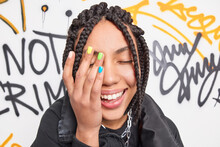 Close Up Shot Of Happy Teenage Girl Makes Face Palm Smiles Broadly Has Colorful Manicure And Dreadlocks Expresses Positive Emotions Poses Against Drawn Graffiti Wall Dressed In Fashionable Clothes