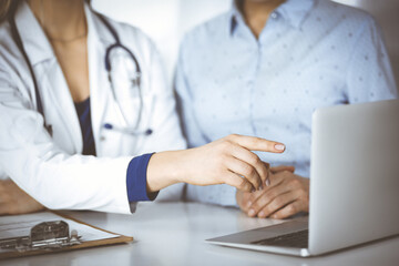 Wall Mural - Unknown woman- doctor and her patient are discussing patient's blood test, while sitting together at the desk in the cabinet in a clinic. Female physician, with a stethoscope, is using a laptop