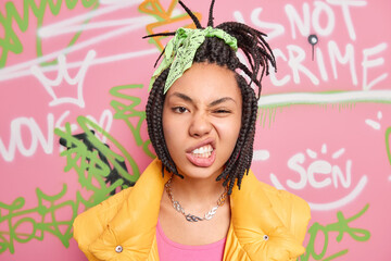 Poster - Teenage lifestyle concept. Cheeky hipster girl with combed dreadlocks clenches teeth smirks face looks self confident at camera spends free time at street poses over graffiti wall background