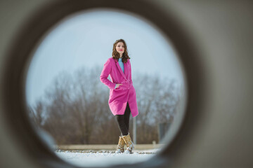 Wall Mural - Full body shot of a young brunette girl through a hole posing in a snowy forest wearing a pink coat