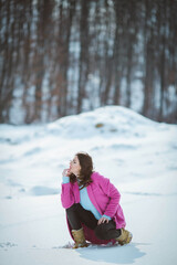 Wall Mural - Young thoughtful brunette girl with eyeglasses  kneeling down in the snow forest wearing a pink coat