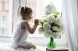 © alexkich - A little girl is sitting on the windowsill. A bouquet of flowers in a vase by the window and a girl sniffing flowers. A little princess in a white dress with a bouquet of white flowers by the window.