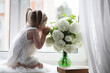 © alexkich - A little girl is sitting on the windowsill. A bouquet of flowers in a vase by the window and a girl sniffing flowers. A little princess in a white dress with a bouquet of white flowers by the window.