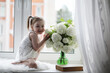 © alexkich - A little girl is sitting on the windowsill. A bouquet of flowers in a vase by the window and a girl sniffing flowers. A little princess in a white dress with a bouquet of white flowers by the window.