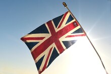 Distressed Old UK Great Britain Flag Against The Sunny Sky. Symbol Of United Kingdom. Weathered British Union Jack. Perfect Stock Photo For Illustrating Negative News About Royal Family