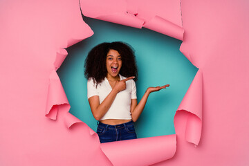 Wall Mural - Young African American woman in torn paper isolated on blue background excited holding a copy space on palm.