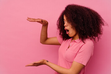 Wall Mural - Young african american woman isolated on pink background shocked and amazed holding a copy space between hands.