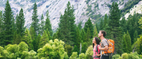 Wall Mural - Hikers hiking in outdoor mountain nature landscape looking away to the side copy space on green pine trees forest. Backpacking tourists woman and man couple with backpacks walking outside banner.
