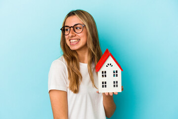 Young caucasian woman holding a house model isolated on blue background looks aside smiling, cheerful and pleasant.