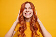 Beautiful carefree teenage girl smiles broadly enjoys favorite music in stereo wireless headphones has long ginger hair spends free time on enjoying playlist isolated over yellow background.