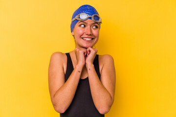 Young swimmer venezuelan woman isolated on yellow background keeps hands under chin, is looking happily aside.