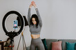 © shevtsovy - Attractive Woman virtual workout at home using smartphone. Fitness workout exercises online woman at home. Fitness influencer personal trainer coach recording on smartphone camera new content for